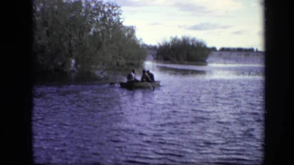Wide 1971 Moscow home-movie view of a small rowboat carrying three men across a broad, tree-edged waterway.