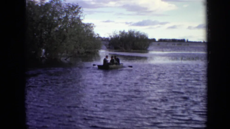 Wide 8mm frame of three men in a rowboat on a Moscow river, with trees to the left and open water behind them.