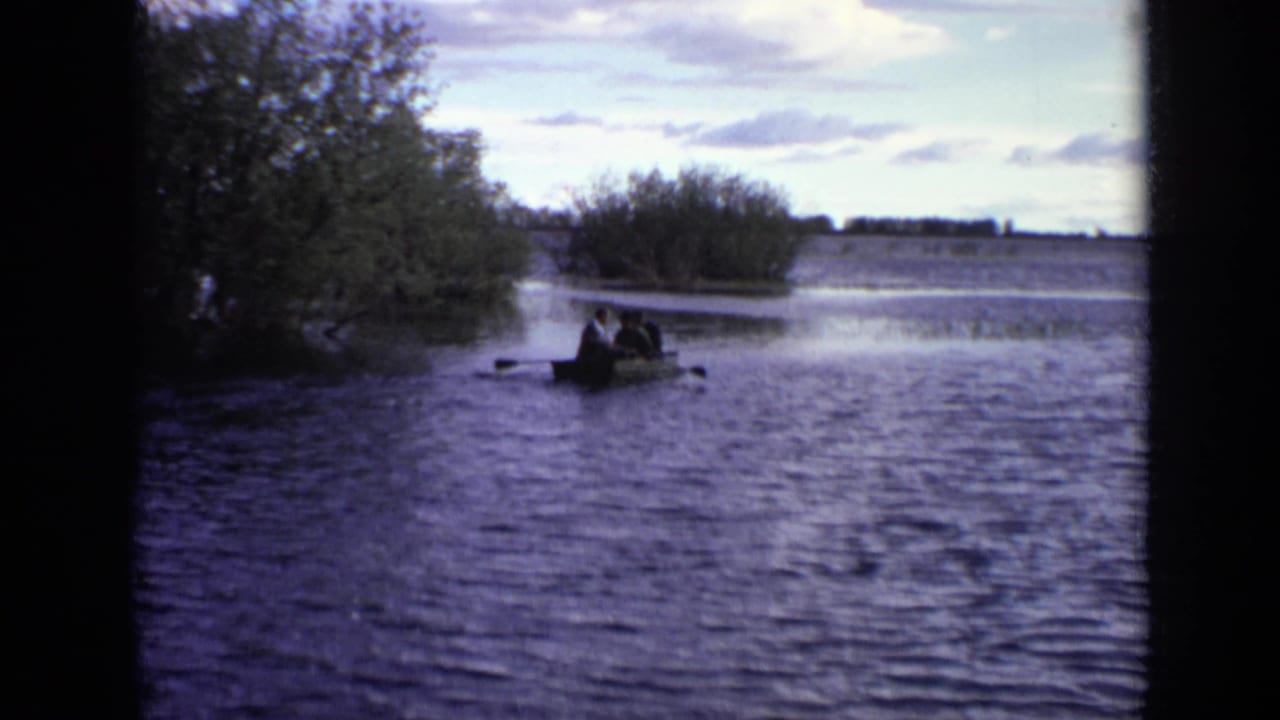 Small rowboat with people near a tree-lined lake shore under a cloudy sky.
