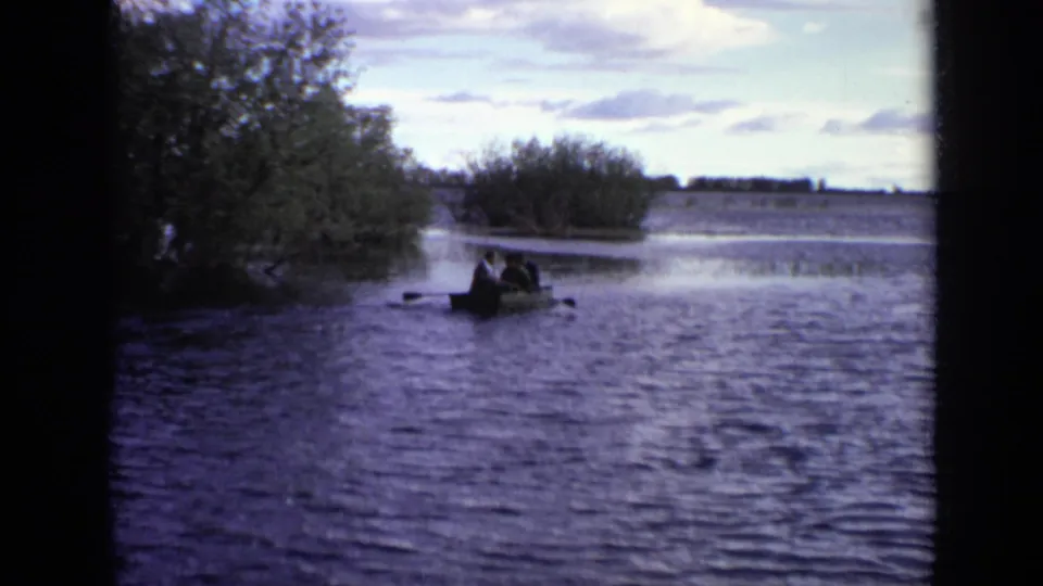 1971 Moscow home-movie frame showing several distant people in a rowboat moving across a lake near trees.