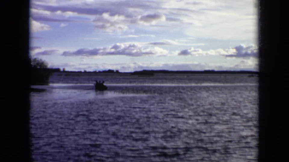 A distant rowboat crosses open lake water in the 1971 Moscow home-movie scene, with a low shoreline beyond.