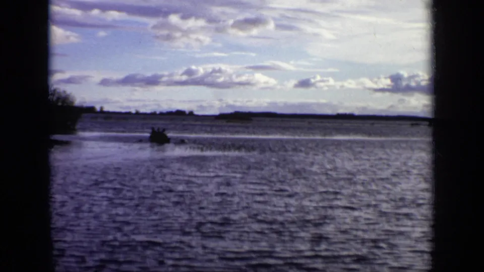 The rowboat is seen farther from shore on a Moscow lake, surrounded by rippled water and a partly cloudy sky.
