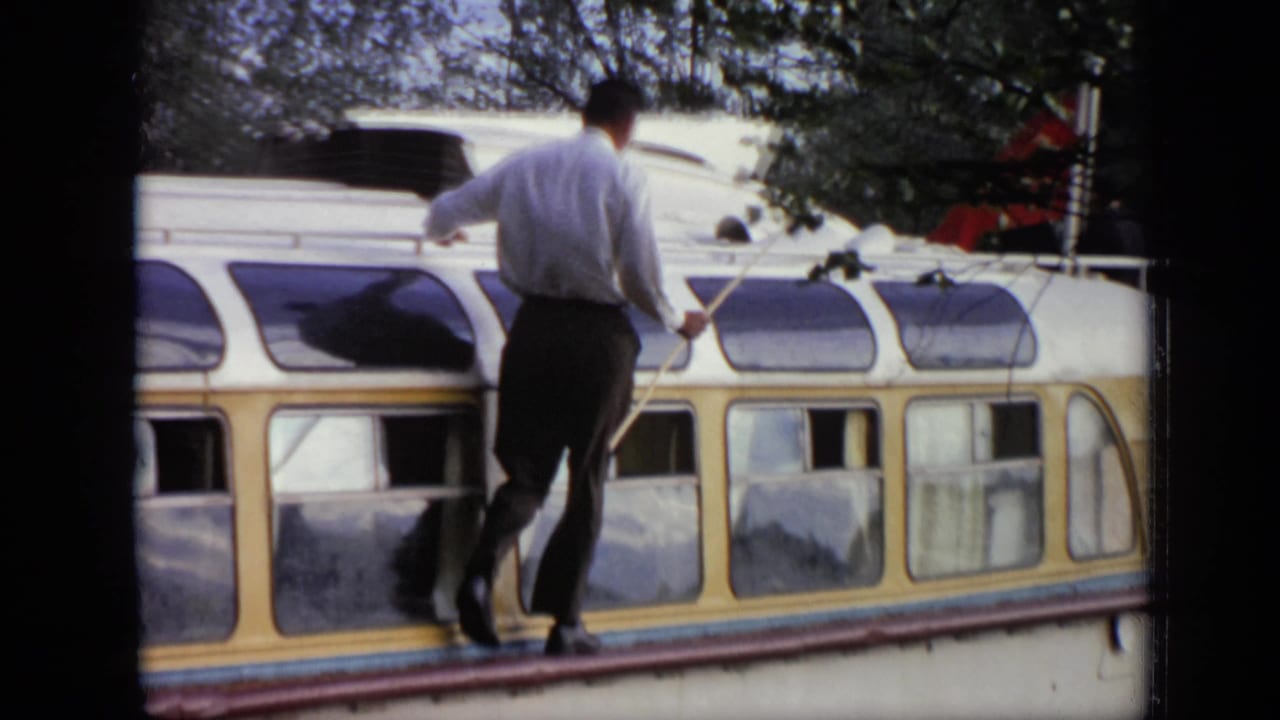 Man holding a long pole while walking on top of a canal boat near trees.