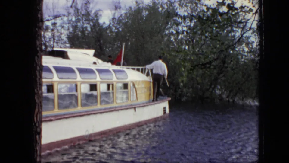 The white and yellow canal boat continues along the tree-lined Moscow waterway, with a man on the roof and a red flag visible.