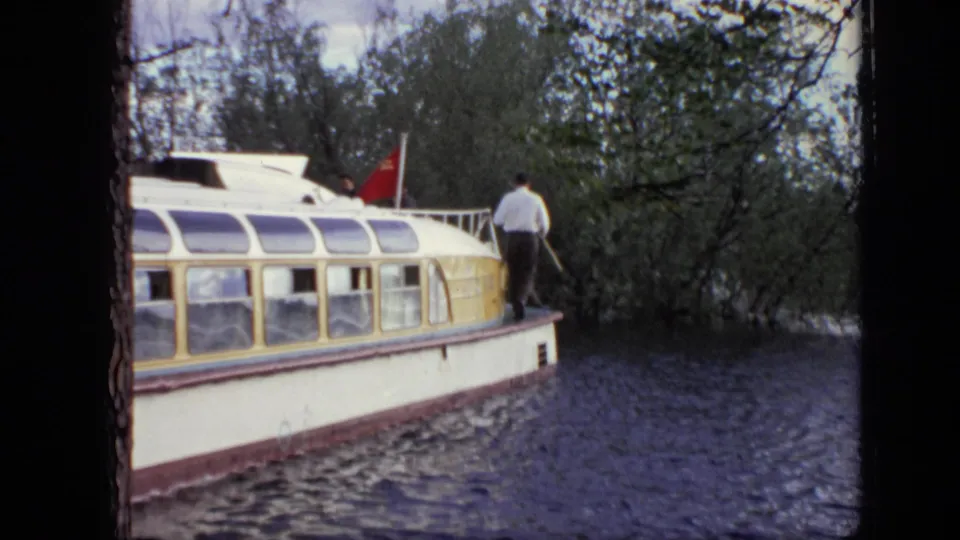 1971 Moscow home-movie frame showing a white and yellow tour boat on rippled water with a man standing near the bow.