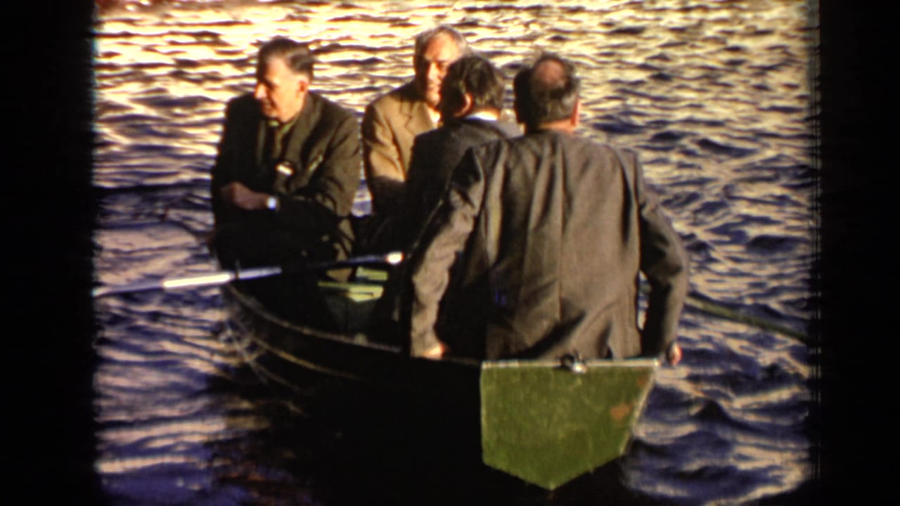 Four men in suits sit in a small rowboat on sunlit river water.