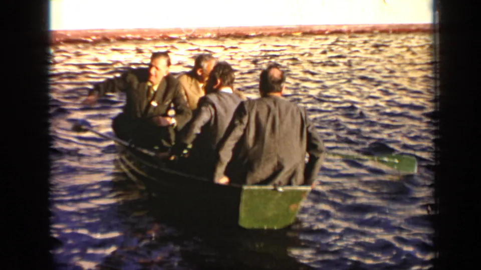 1971 Moscow home-movie frame showing suited men in a rowboat, viewed from behind as the boat moves across the river.