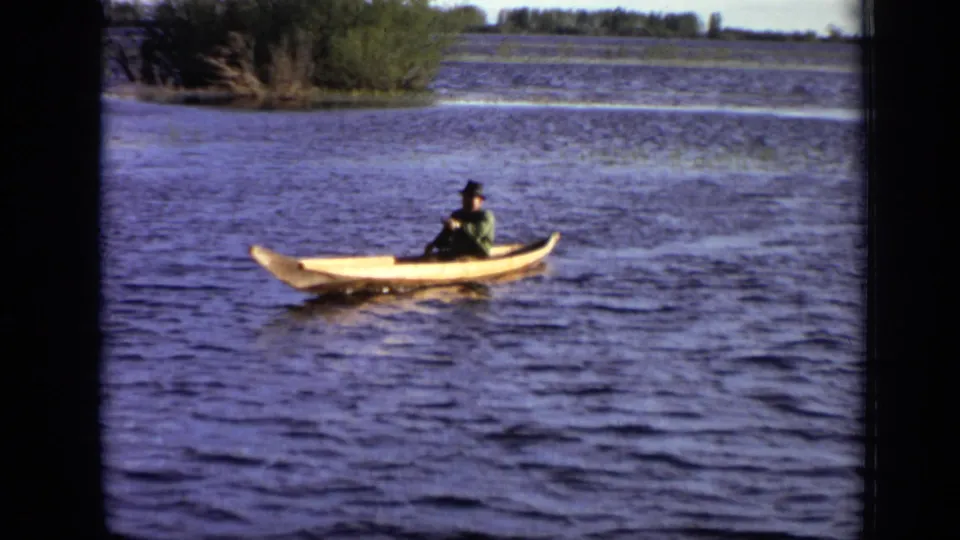1971 Moscow home-movie frame of a lone hatted paddler in a light wooden kayak on open river water.