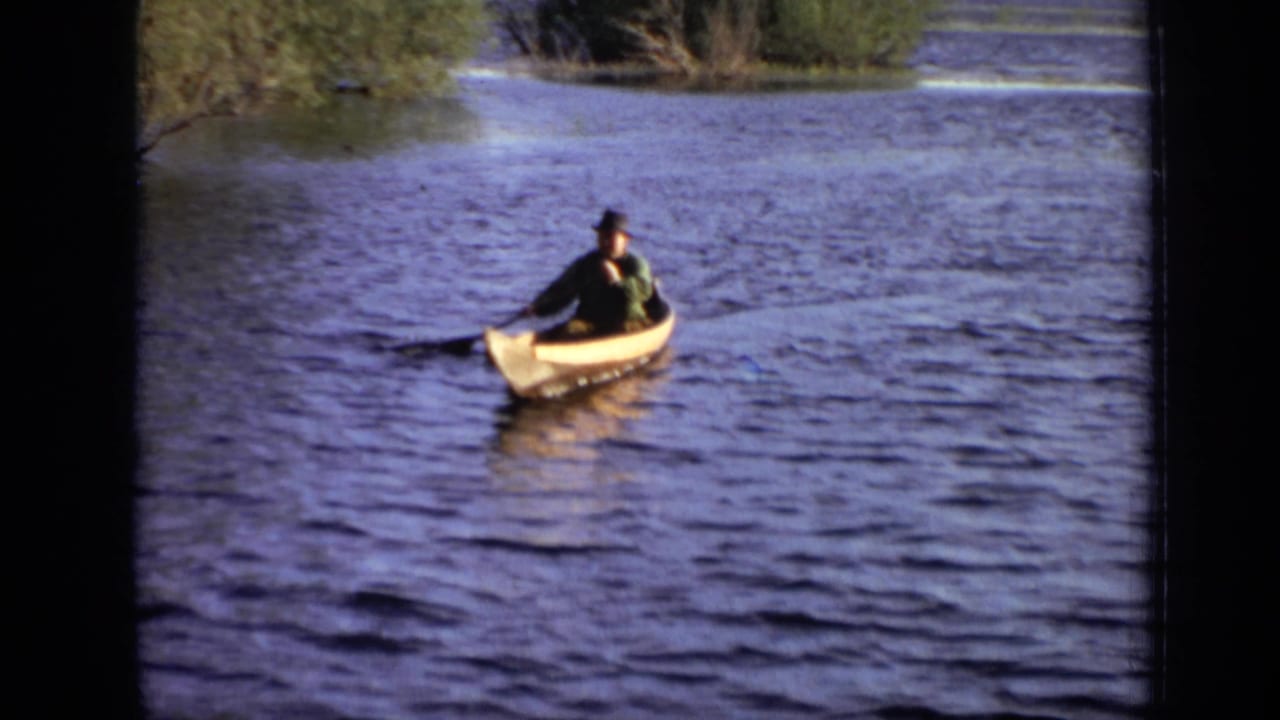In metadata-supported 1971 Moscow footage, a man in a dark hat paddles a yellow canoe across open water with shoreline trees behind him.