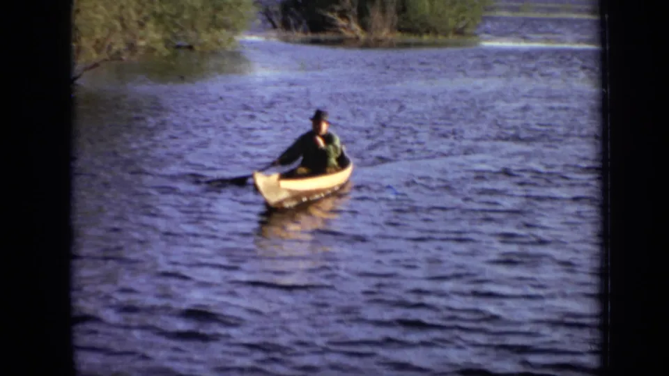 In metadata-supported 1971 Moscow footage, a man in a dark hat paddles a yellow canoe across open water with shoreline trees behind him.
