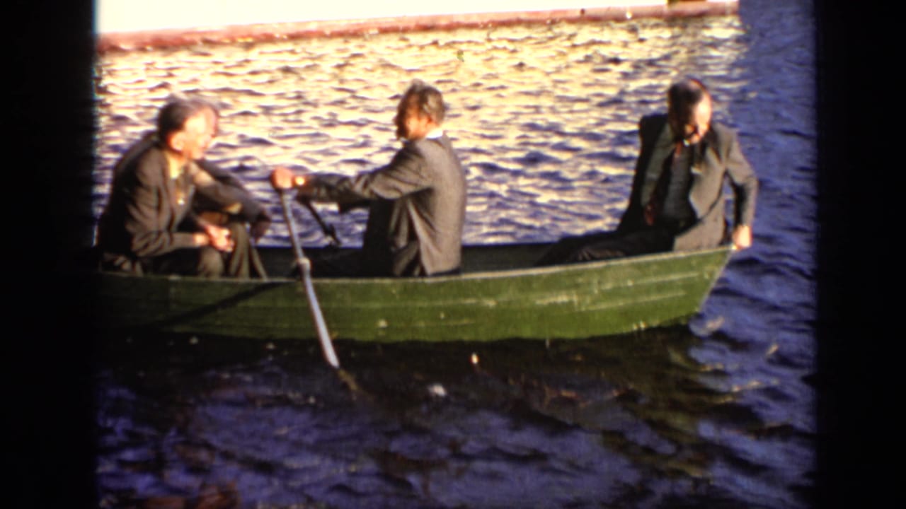 Three adult men in suits sit in a green rowboat on sunlit water, with one handling an oar during the Moscow boating scene.