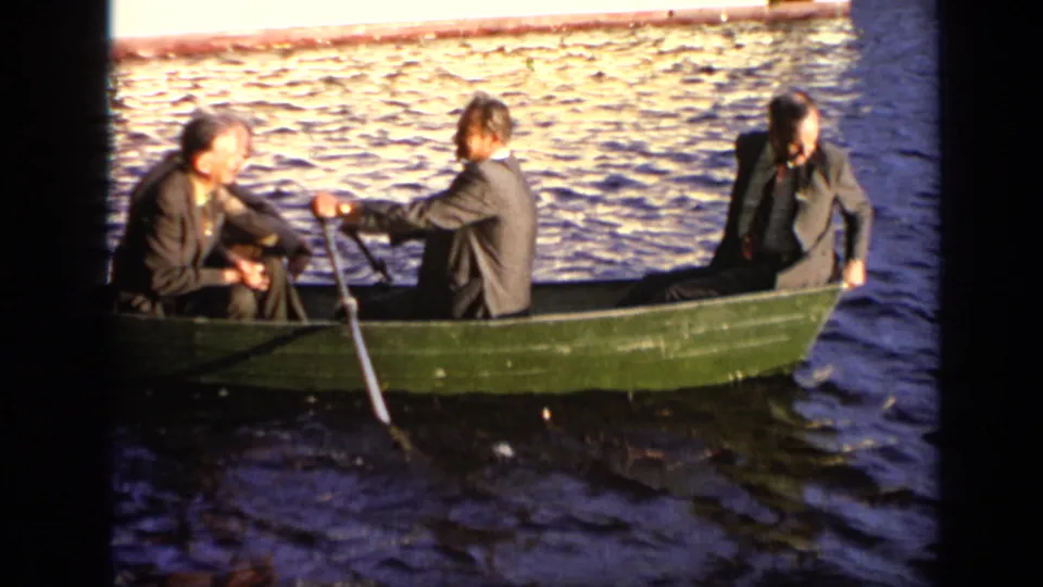 Three adult men in suits sit in a green rowboat on sunlit water, with one handling an oar during the Moscow boating scene.