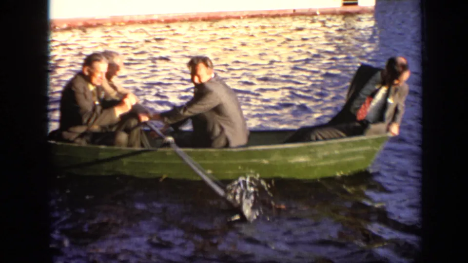 A closer 8mm view shows suited older men in the green rowboat as an oar pulls through the water and splashes beside the hull.