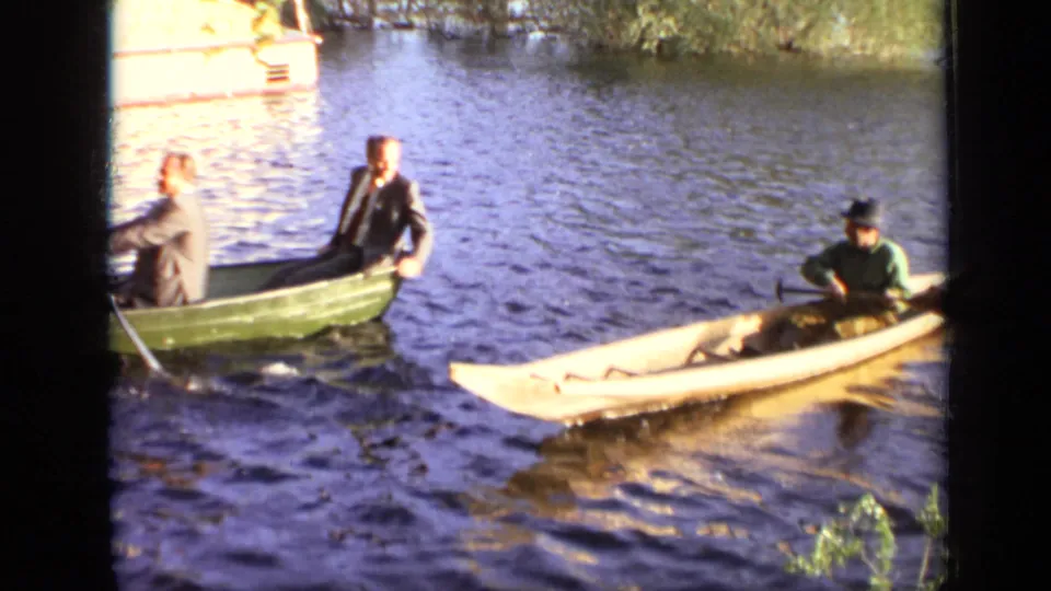 The green rowboat with suited men moves near the yellow canoe paddled by a man in a hat, both crossing the same sunlit waterway.