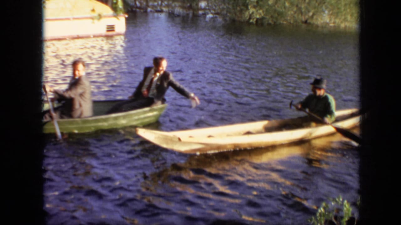 Two suited young men in a green rowboat beside a pale canoe on rippled water.