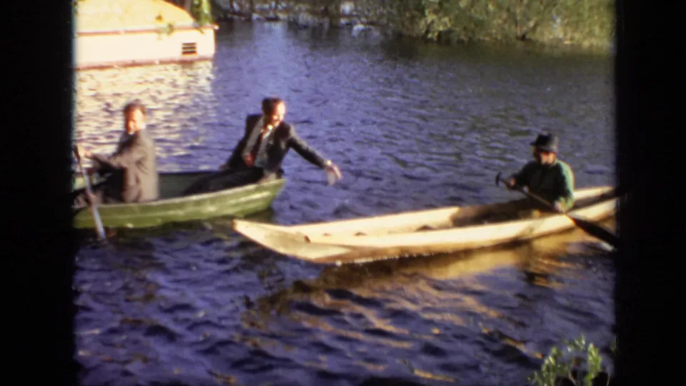1971 Moscow home-movie frame of suited young men maneuvering a green rowboat beside a pale canoe on a river.