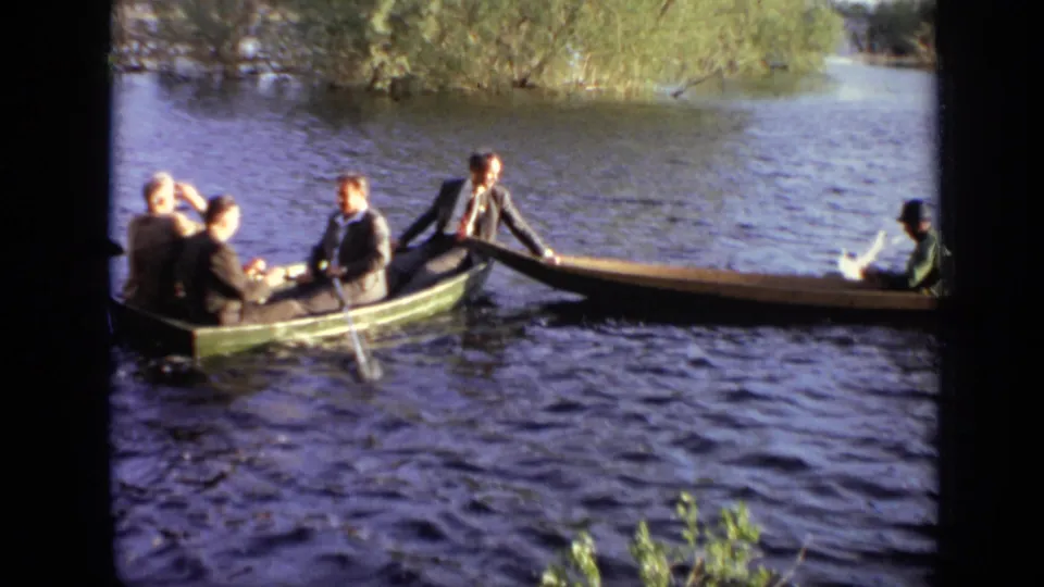 A group of young men in suits crowd into a green rowboat beside a canoe on a Moscow waterway.