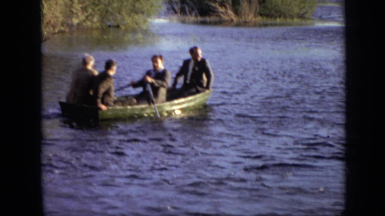 Four people sit in a small green rowboat on rippled water near reeds.