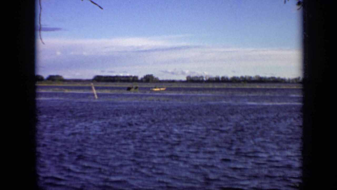 Wide river view with distant boats, grasses, tree line, and dark 8mm frame edges.