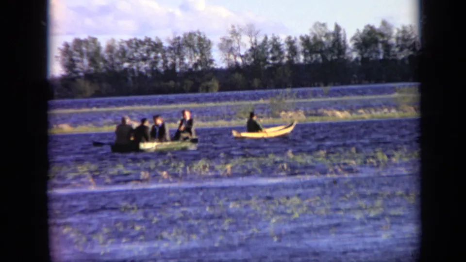 Final 1971 Moscow 8mm frame with men in a green rowboat, a lone canoeist nearby, rippled water, grasses, and a tree-lined bank.
