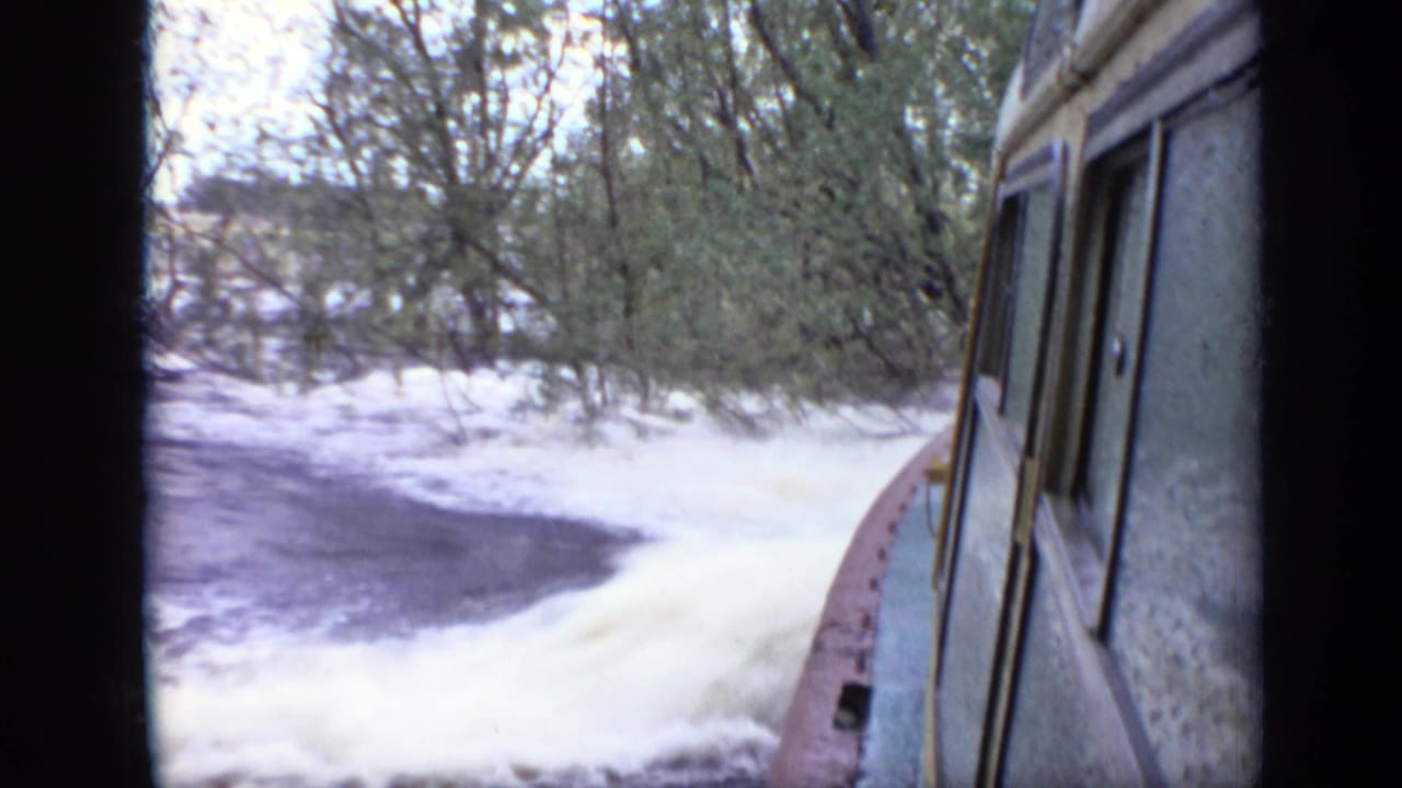Cabin boat side moving past foamy water and trees along a riverbank.