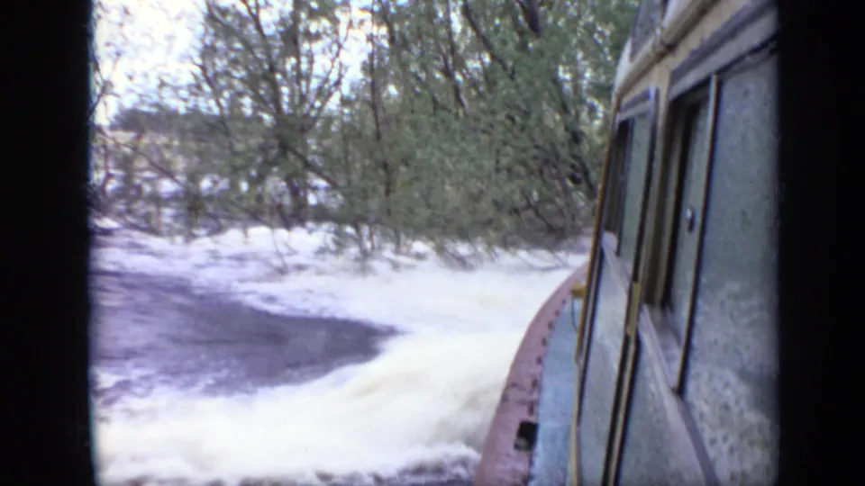 1971 Moscow 8mm frame from a moving cabin boat beside foamy wake and a wooded shoreline.