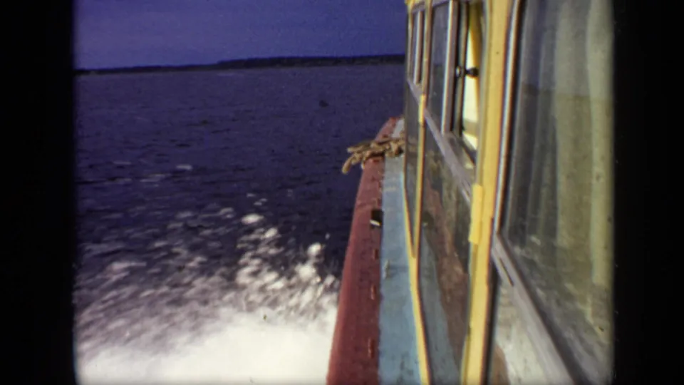 Handheld 1971 boat-travel frame showing the vessel side, churning wake, dark blue water, and distant wooded shoreline in Moscow-area footage.