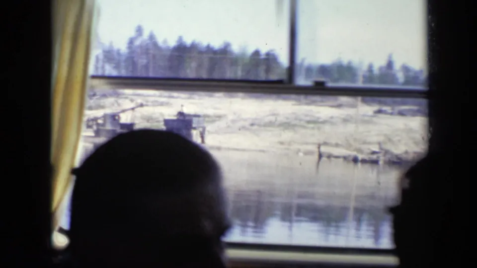 Interior-facing 8mm frame from the boat with a passenger silhouette near the window and a pale shoreline, water, and small dock structures outside.