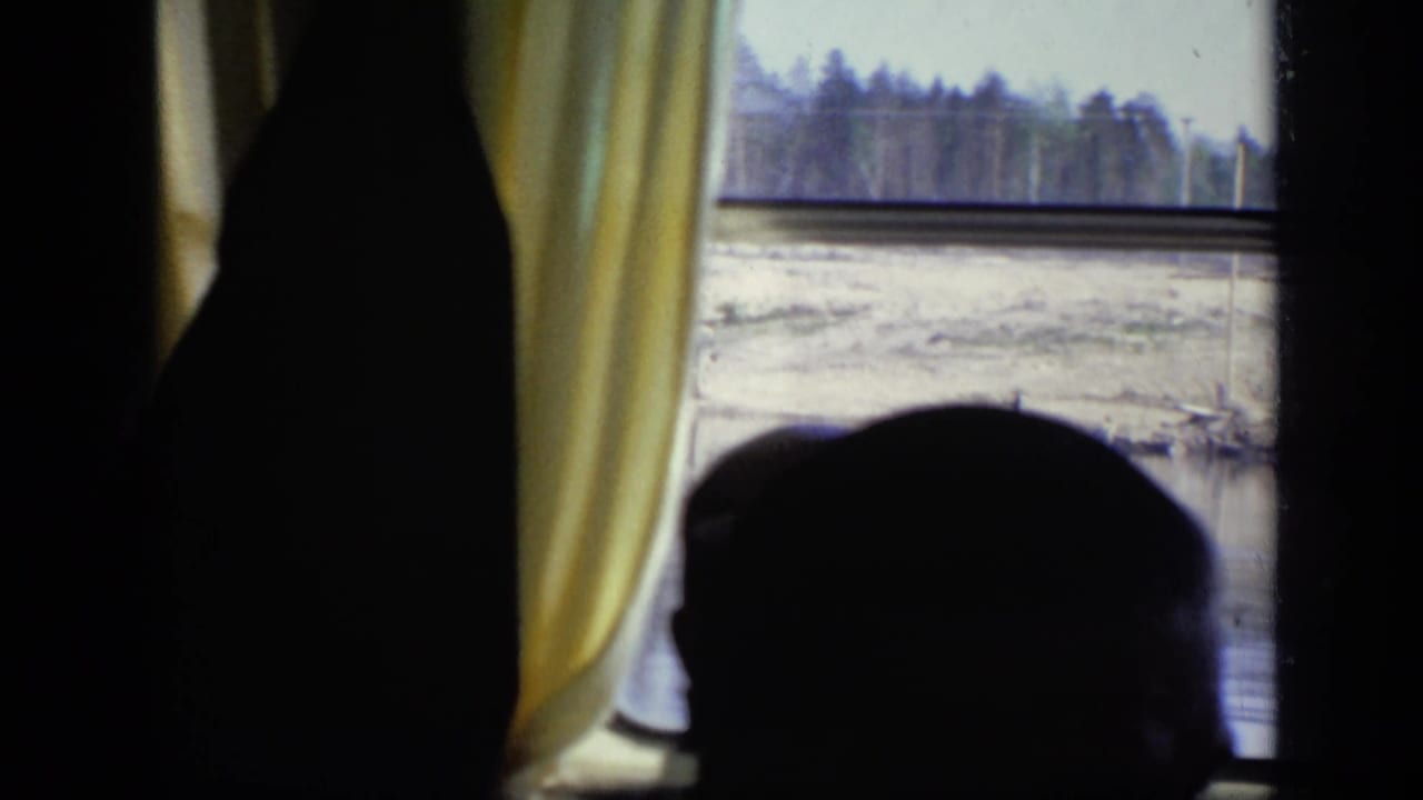 1971 Moscow home-movie frame showing a dark passenger silhouette against yellow curtains and a riverside view through a window.