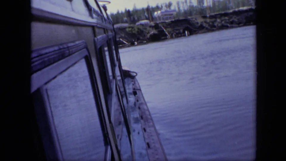 1971 Moscow frame looking along a vehicle or vessel exterior beside water, with a wooded bank and small structures beyond.