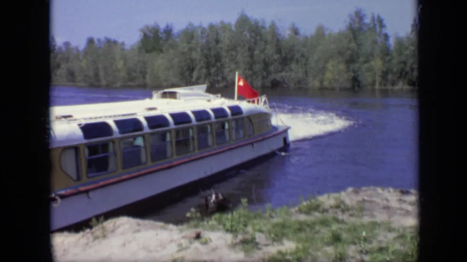 1971 Moscow 8mm frame of a white passenger boat near shore, with a red flag, broad windows, water, and trees visible.