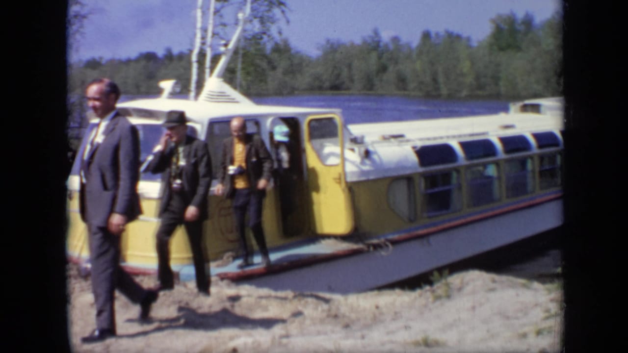 Suited men step away from a yellow and white boat on a sandy shore.