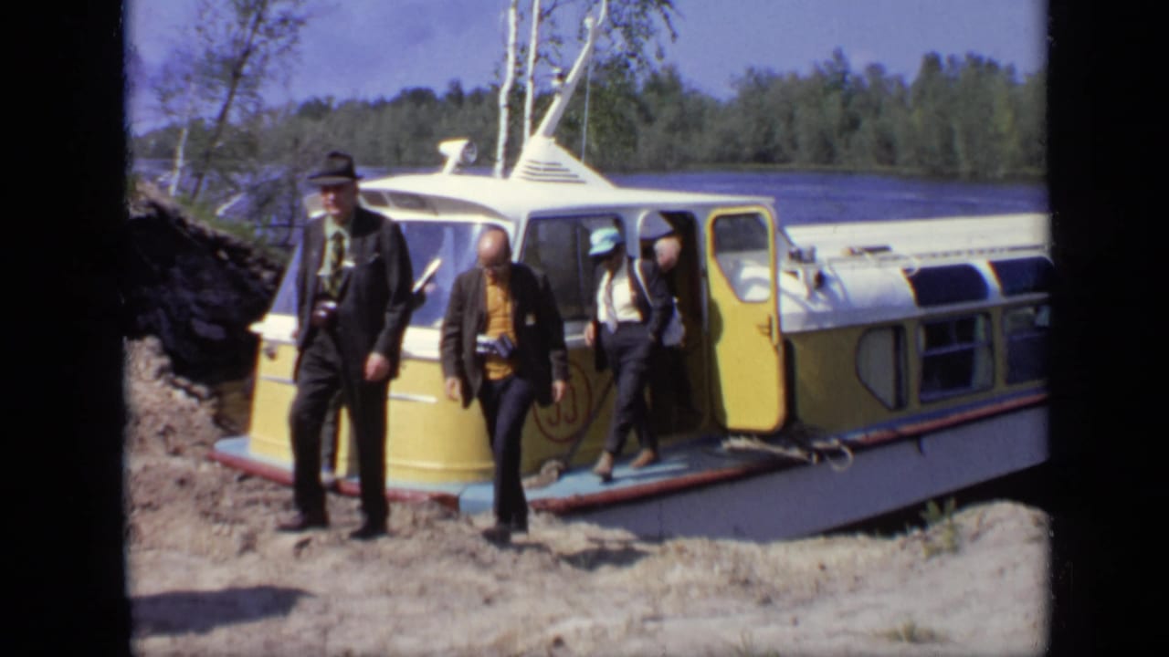 Several formally dressed men, some carrying cameras, stand beside an open yellow boat door near water in the 1971 Moscow footage.