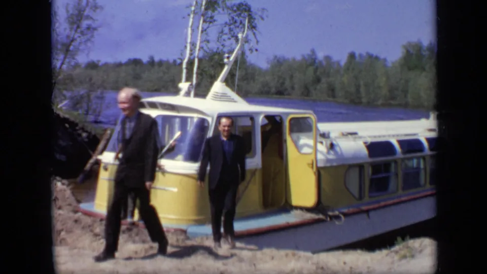 Two men in suits walk across the sandy shore in front of the yellow and white boat, with lake and trees behind them.