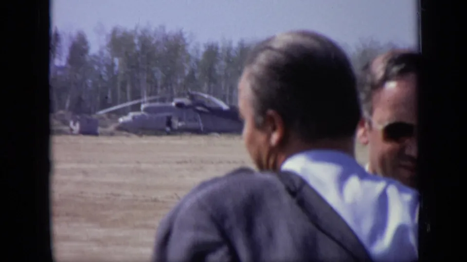 A man seen from behind and another man in sunglasses look across an open field toward a helicopter in the 1971 Moscow footage.