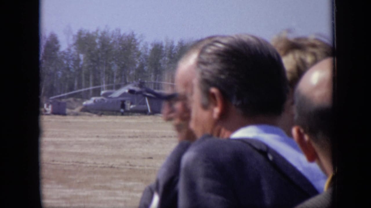 1971 Moscow 8mm frame showing suited men in the foreground and a parked helicopter across a flat open field.