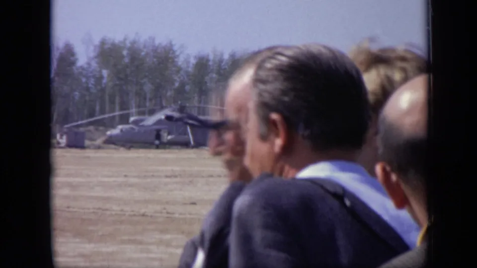 1971 Moscow 8mm frame showing suited men in the foreground and a parked helicopter across a flat open field.