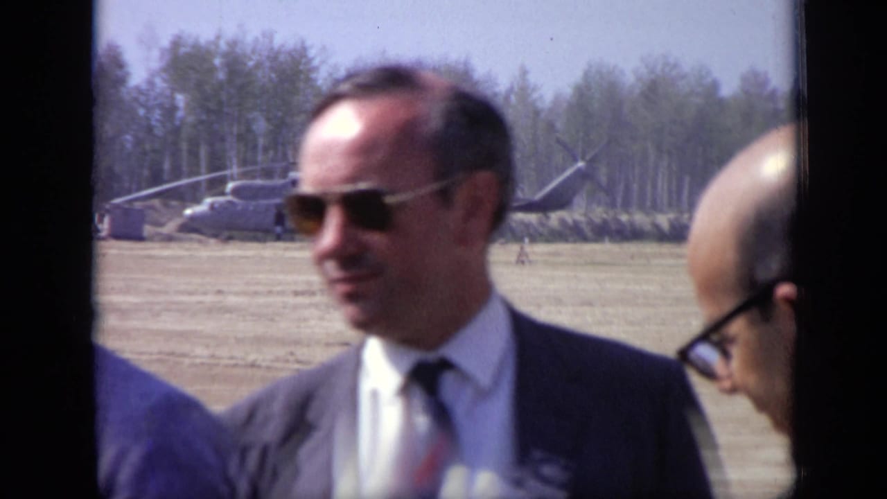 A man in sunglasses and a suit stands near camera with a helicopter and trees behind him.