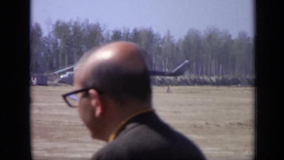1971 Moscow 8mm frame with a man in glasses passing close to lens and a helicopter parked beyond the open field.