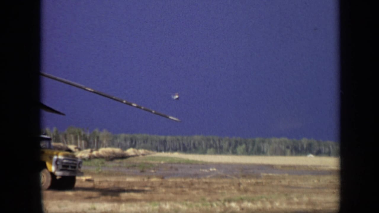 Yellow utility truck beside an open field with a helicopter rotor or boom crossing the sky.