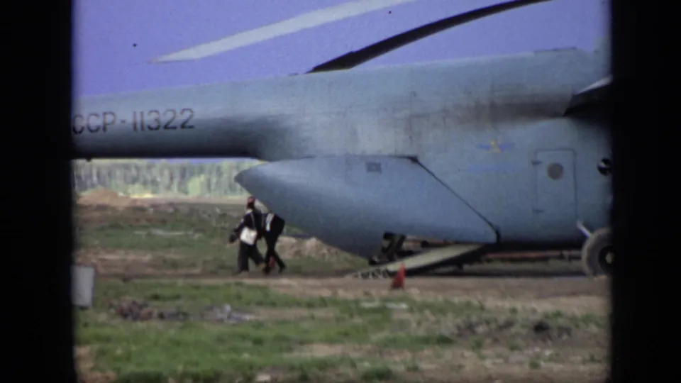 Two adult men in dark suits walk near the Soviet-marked helicopter in the 1971 Moscow-area rural field scene.