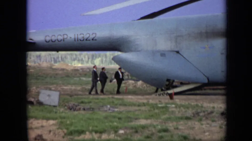Three adult men in dark suits gather near the Soviet-marked helicopter and ramp area in 1971 Moscow-area 8mm footage.