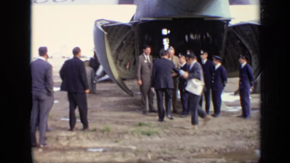 A suited man moves away from the open aircraft ramp while officers and other men remain gathered on the Moscow airfield in 1971 footage.