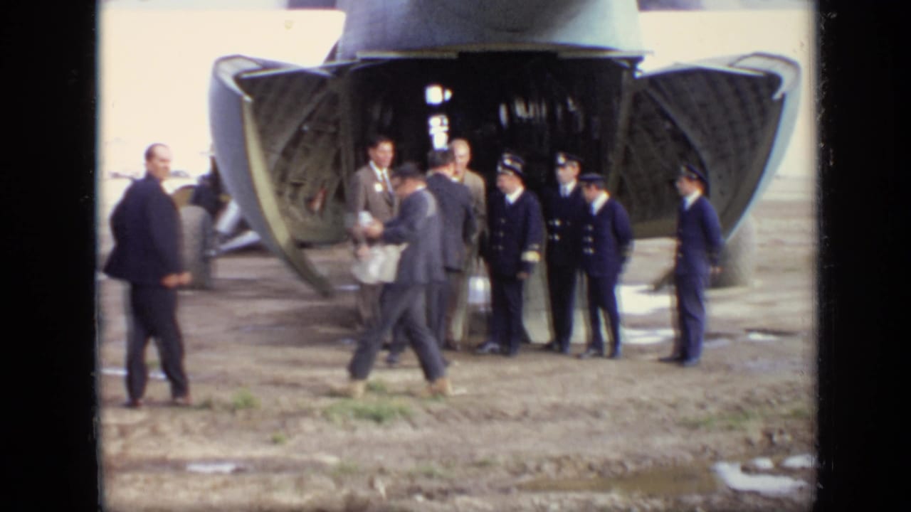 Men stand near the open rear ramp of a green cargo aircraft on an airfield.