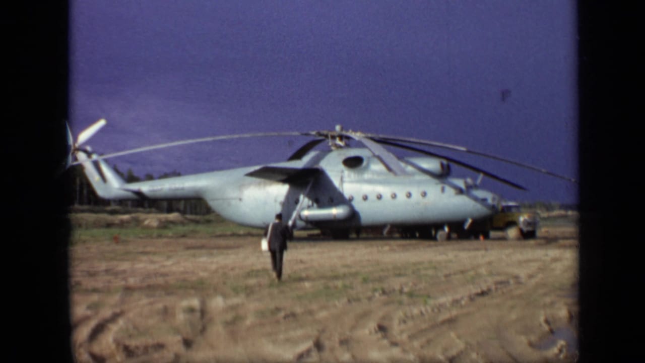 Large light-gray helicopter parked on muddy ground with a man walking toward it.