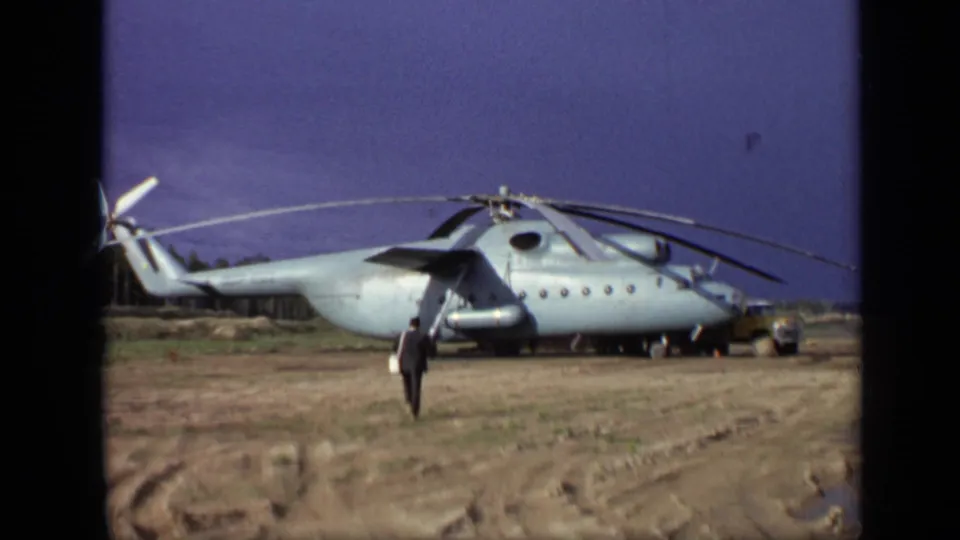 1971 Moscow 8mm frame showing a man crossing a muddy field toward a large light-gray helicopter with rotors at rest.