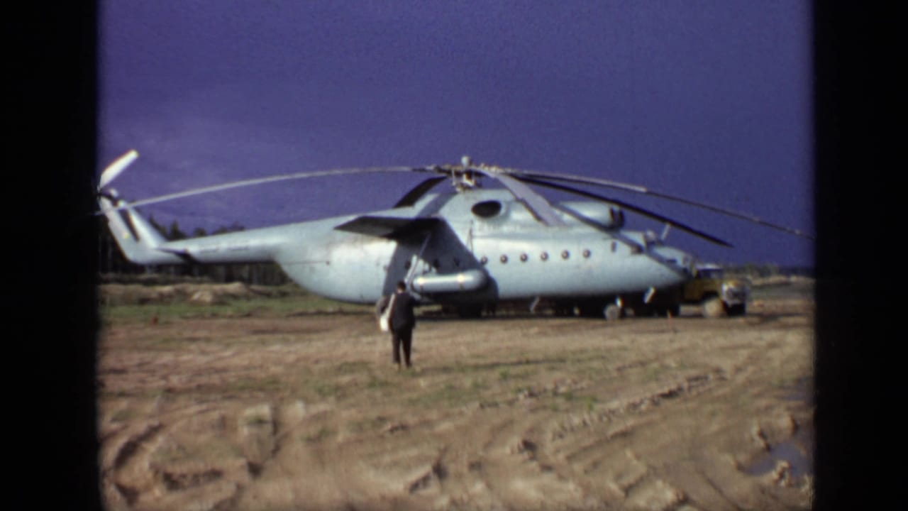 1971 Moscow 8mm frame with a man near the nose area of a large parked helicopter and a small yellow utility vehicle at right.