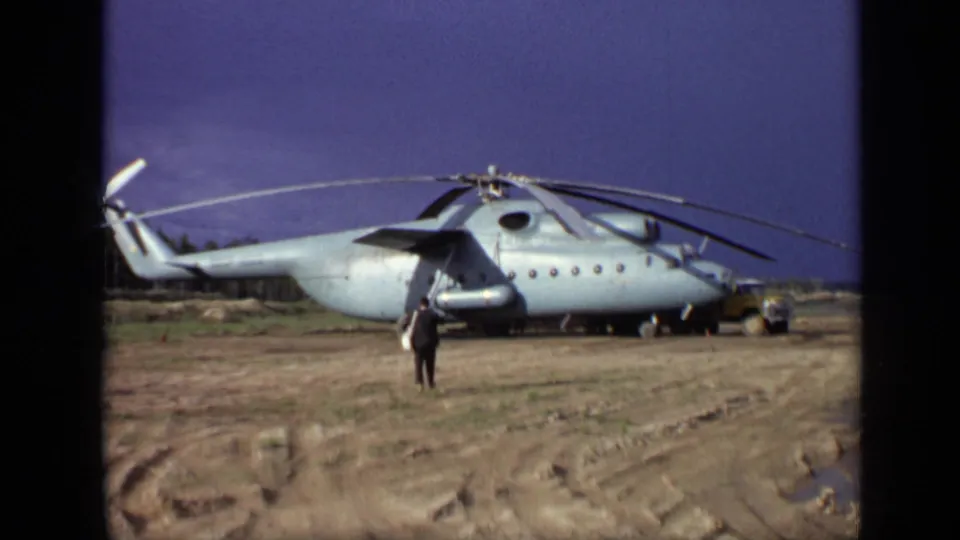 1971 Moscow 8mm frame with a man near the nose area of a large parked helicopter and a small yellow utility vehicle at right.