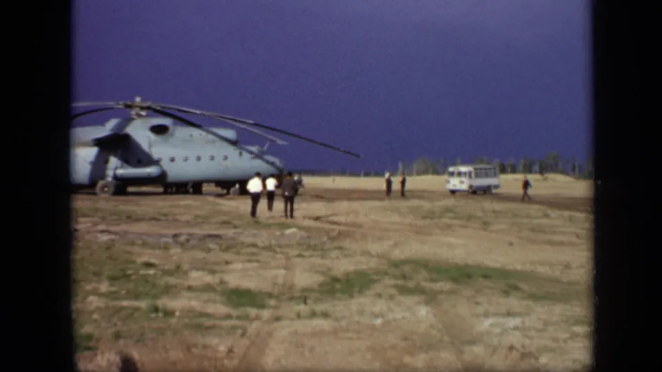 Wide 1971 Moscow 8mm view of a helicopter on a dry grassy field with adults walking and a white van parked to the right.