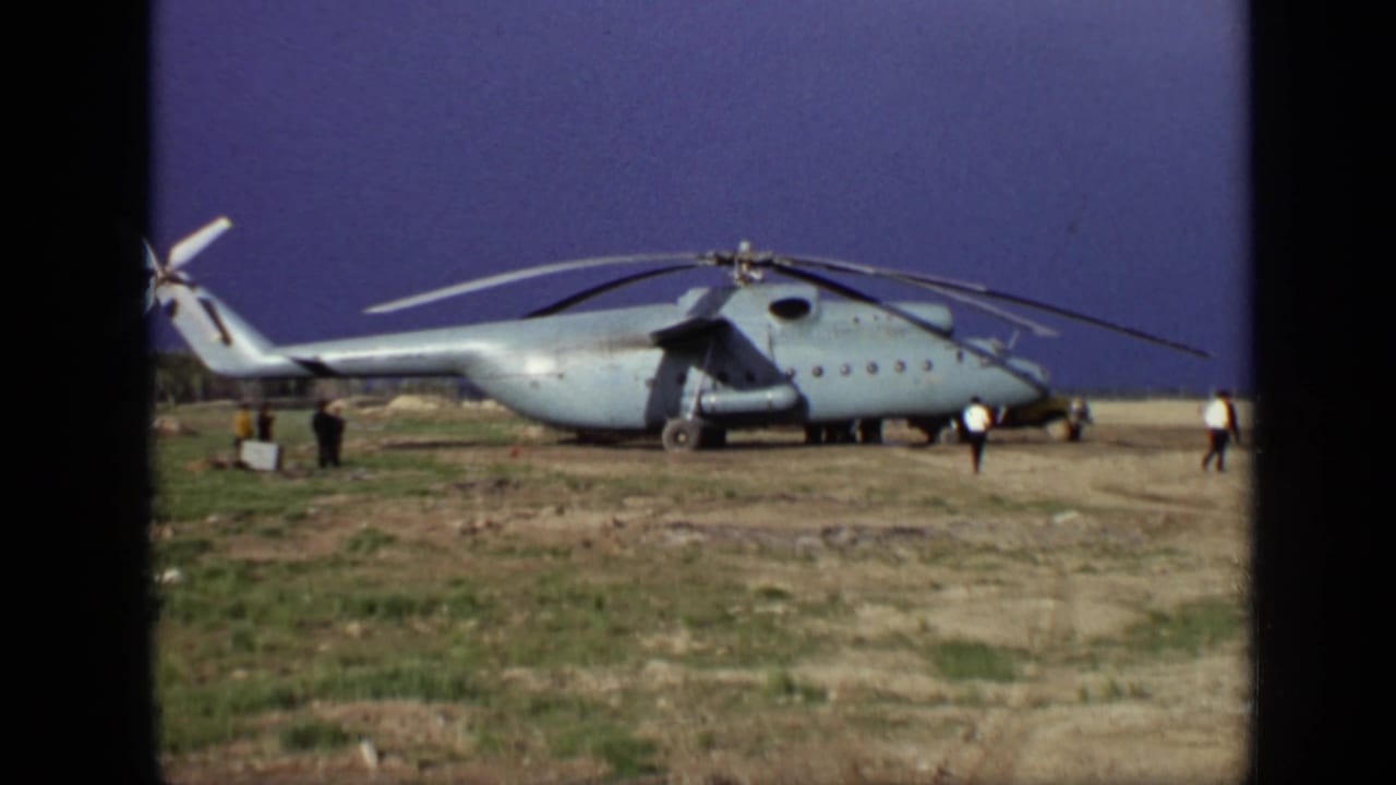 1971 Moscow 8mm frame showing a large parked helicopter with rotors still and several distant people on rough open ground.